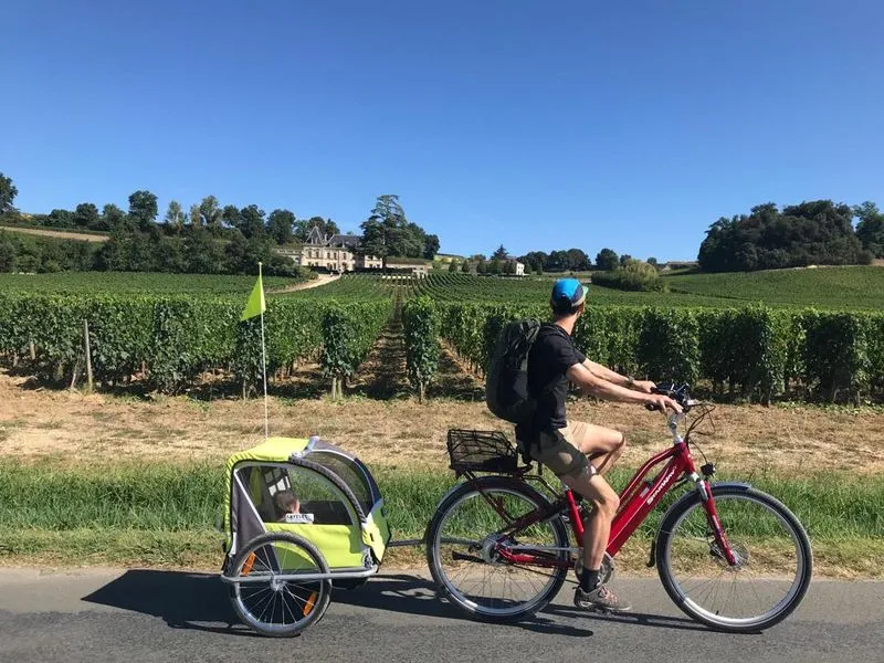 Cyclist on a red bike with a yellow-green trailer on a road alongside Bordeaux vineyards, with a wine château visible in the background on a sunny day.