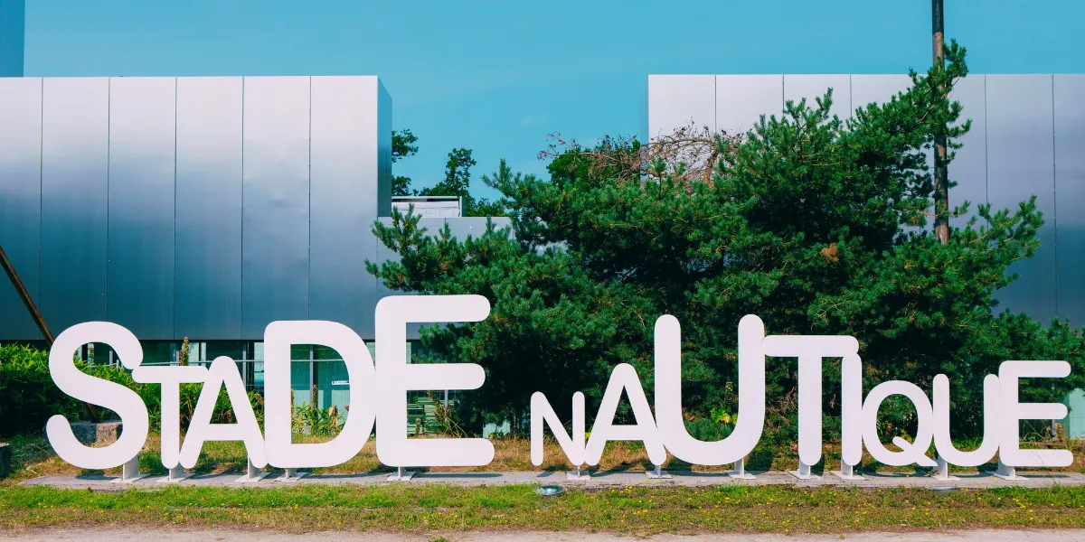 White letter sign reading « STADE NAUTIQUE » in front of a modern building with blue and white facades and lush green vegetation in Bordeaux.