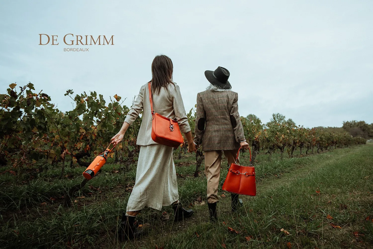 Two stylish women walk through Bordeaux vineyards, carrying red accessories and a wine bottle, embodying the luxury and wine heritage of the region.