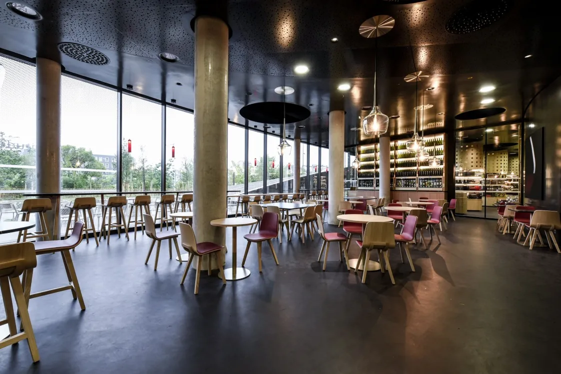 Modern interior of Latitude 20 wine bar in Bordeaux featuring high tables, wooden and velvet chairs, wine bottle display behind the bar, black ceiling with pendant lights, and expansive city views through floor-to-ceiling windows.