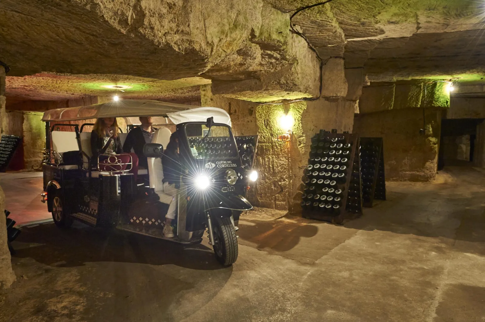 An illuminated tuk-tuk tourist vehicle in the Cordeliers underground catacombs in Bordeaux, carrying passengers with rows of wine bottles stored in the vaulted galleries.