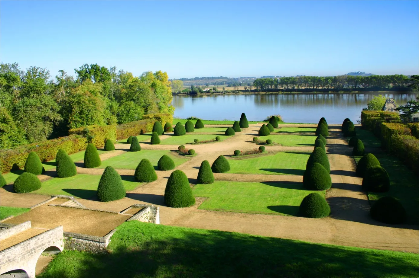 Formal French gardens at Château de Vayres featuring cone-shaped boxwood topiaries, manicured lawns, gravel pathways, and the Dordogne river in the background, near Bordeaux.