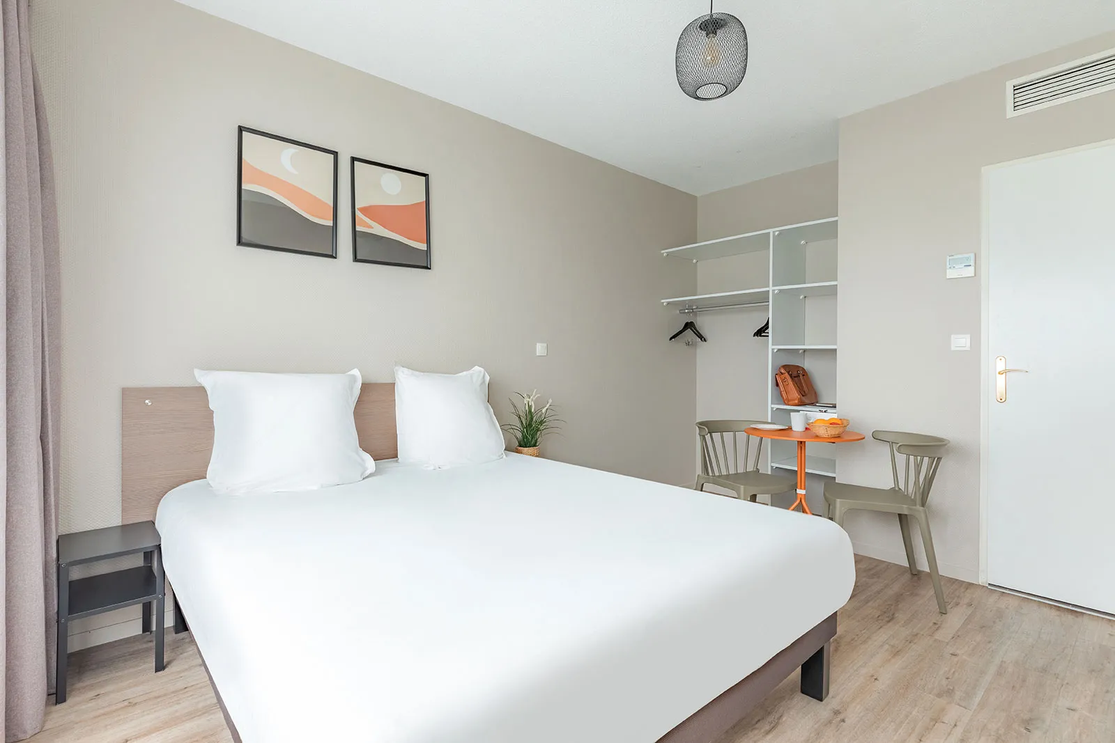 Modern bedroom in a Bordeaux apartment featuring a double bed with white linens, an orange desk, grey chairs, built-in wardrobe, and minimalist décor.
