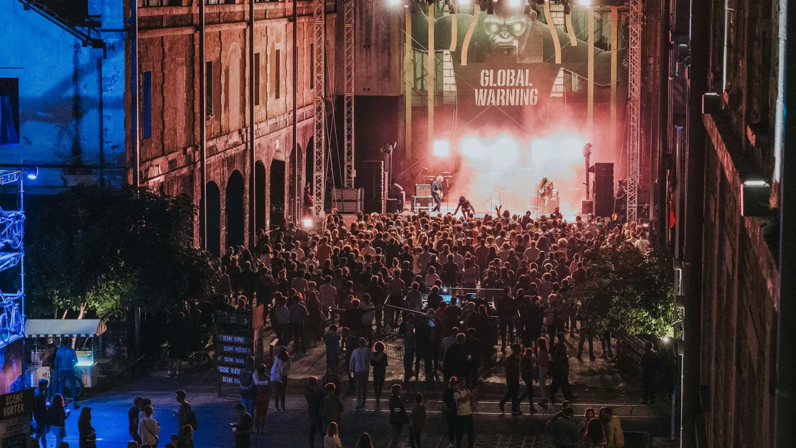 Large crowd of spectators at a night concert in a narrow street in Bordeaux, with an illuminated stage displaying 'GLOBAL WARNING' and red brick buildings on either side