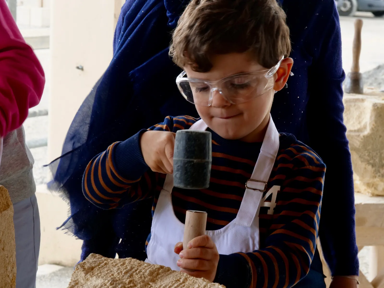 A child wearing safety glasses and a white apron chiseling stone with a chisel and mallet during a medieval workshop in Bordeaux.