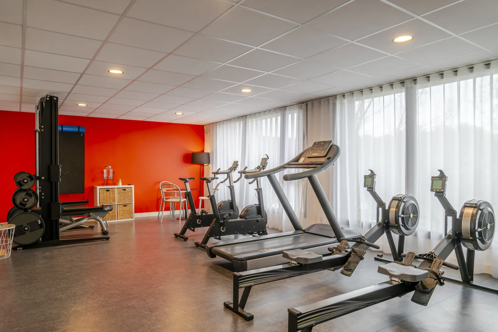 Modern fitness room at Ibis Styles Bordeaux Lac Bruges hotel featuring treadmills, exercise bikes, weightlifting equipment, vibrant orange walls, white ceiling, large windows with curtains and natural light.