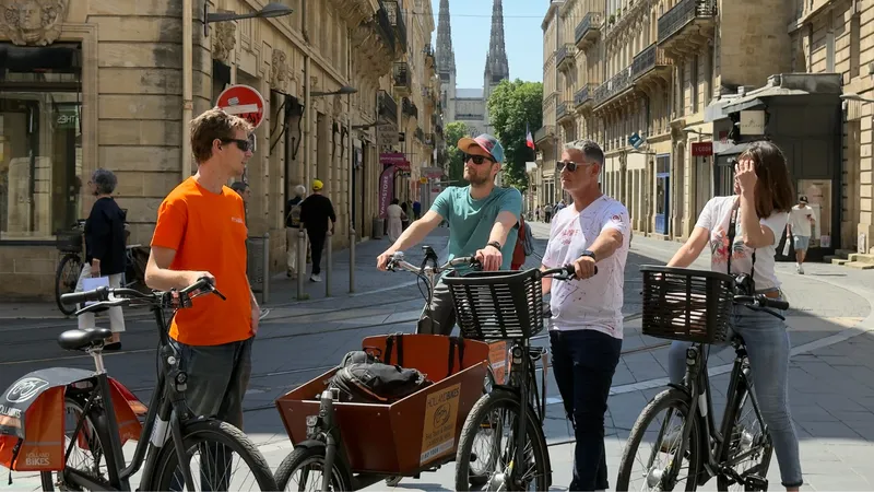 A group of five tourists on bicycles in a paved street in Bordeaux, with historic buildings and a church in the background. Some bikes are equipped with transport baskets.