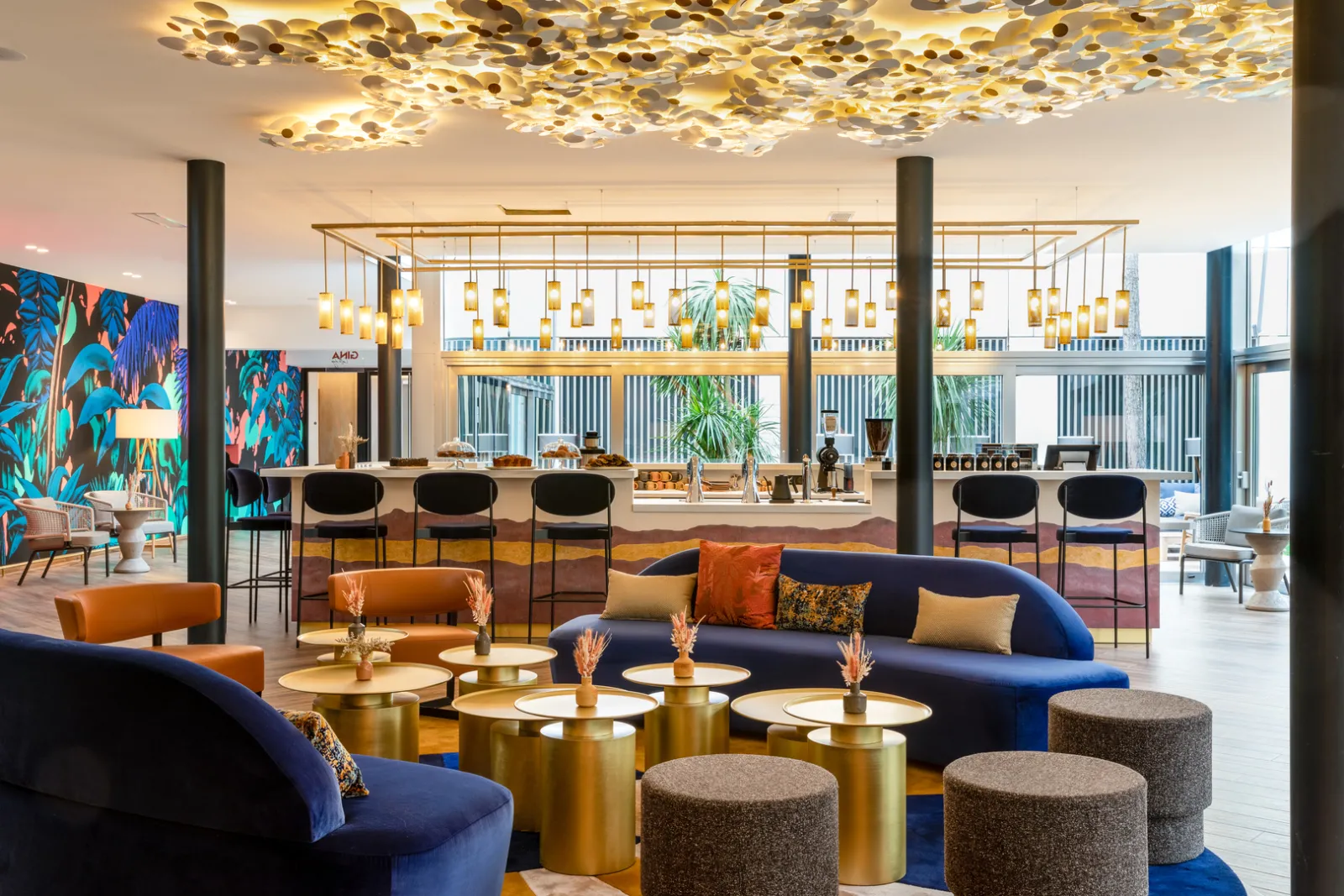 Modern lobby of Renaissance Bordeaux Hotel featuring blue and orange designer furniture, golden ceiling chandelier, bar area in background, and large windows overlooking the city.