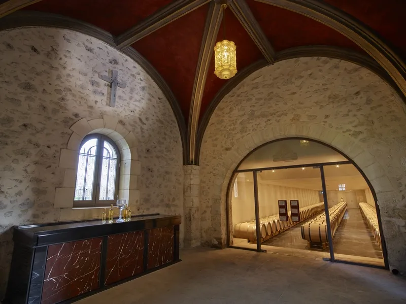 Interior of a chapel at Château Lafaurie Peyraguey in Bordeaux, featuring stone vaults, a wall cross, an arched window, and a glass door leading to a barrel-lined gallery.