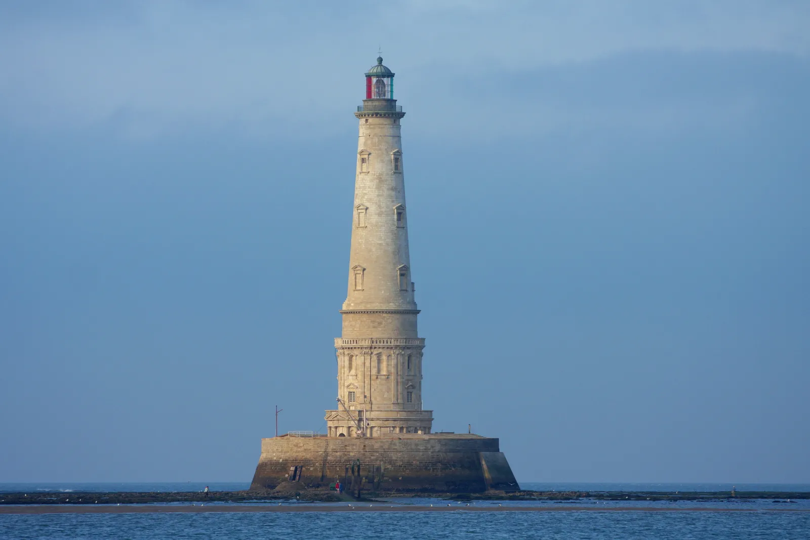Cordouan Lighthouse, a magnificent white stone beacon standing on a rocky island at sea, featuring a red lantern room topped with a spire, against a clear blue sky.