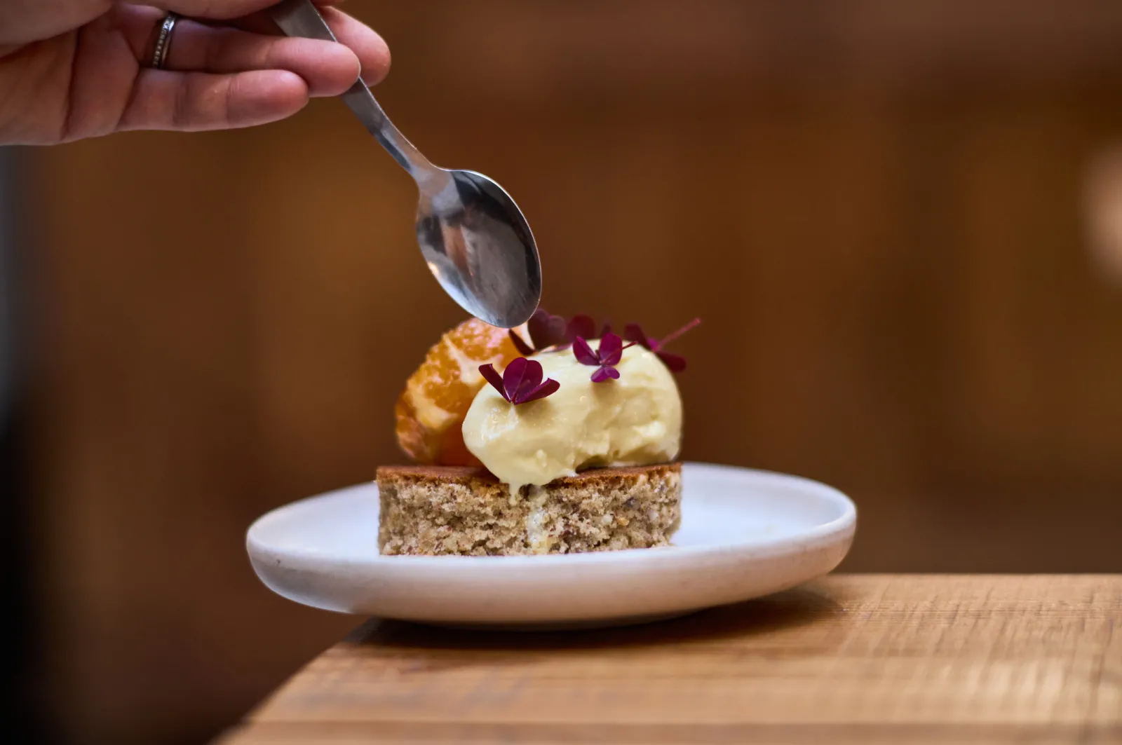 A pastry chef drizzles sauce with a spoon over an elegant dessert featuring a grain biscuit, pale cream, golden pastry, and purple edible flowers.