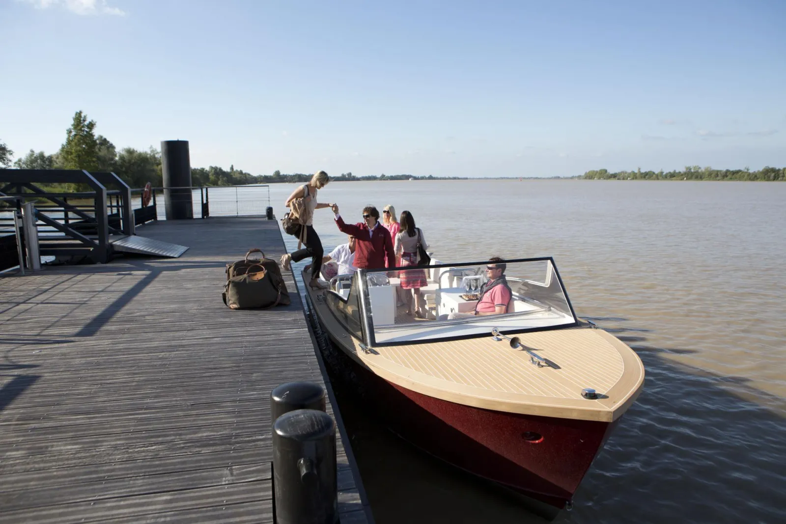 Passengers boarding the Grattequina Water Taxi in Bordeaux, moored at a wooden dock on the Garonne River, with trees and the river visible in the background.