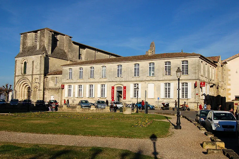 Façade of the Wine House of Saint-Emilion in Bordeaux, stone building with adjoining Romanesque church, green lawn in foreground and parked cars