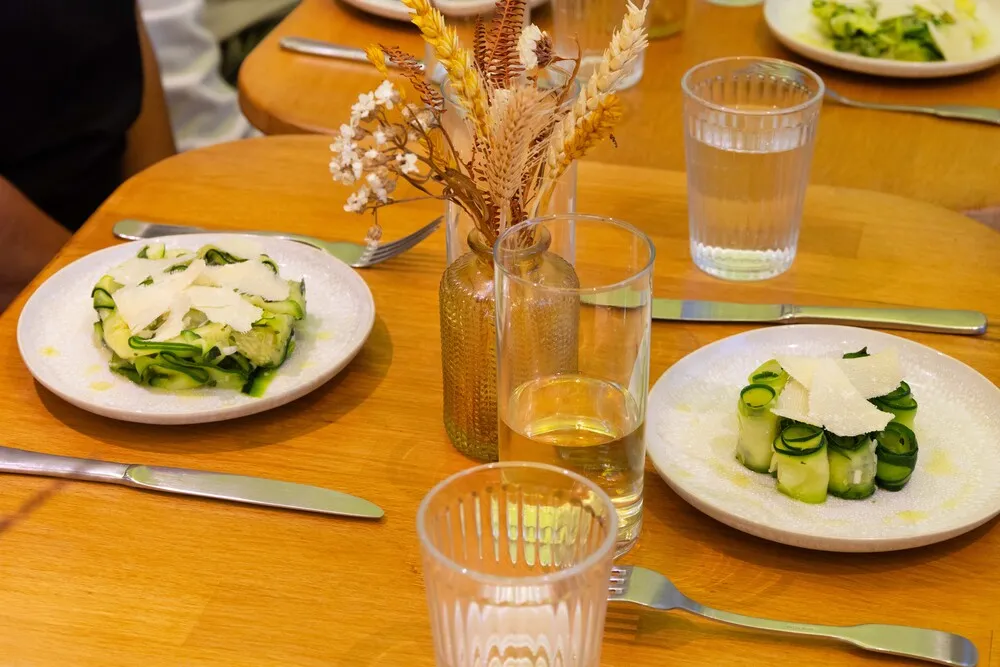 Restaurant table featuring two plates of sliced zucchini with white cheese, water glasses, and a dried flower arrangement in the center