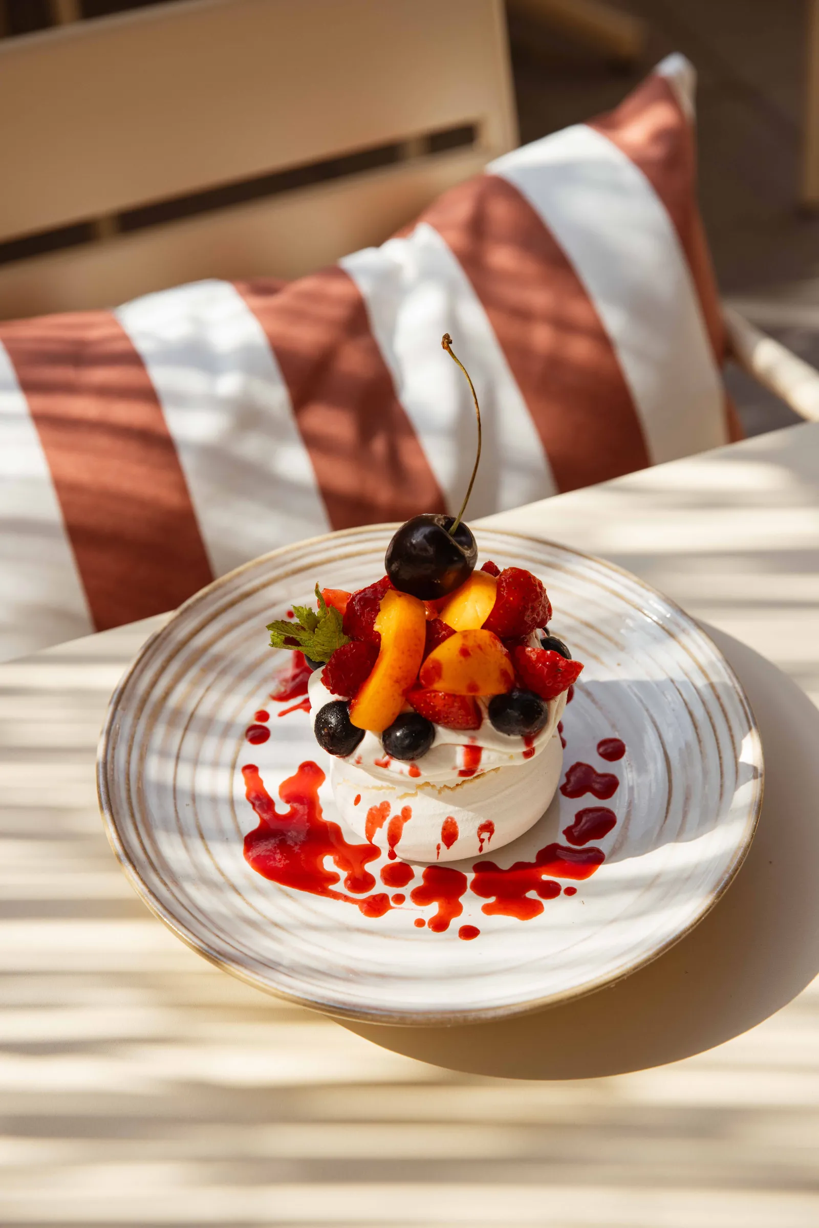 Elegant plated dessert featuring layered white cream, red and yellow fruits with a black cherry on top, decorated with red sauce drizzle on a white plate, with striped cushion background