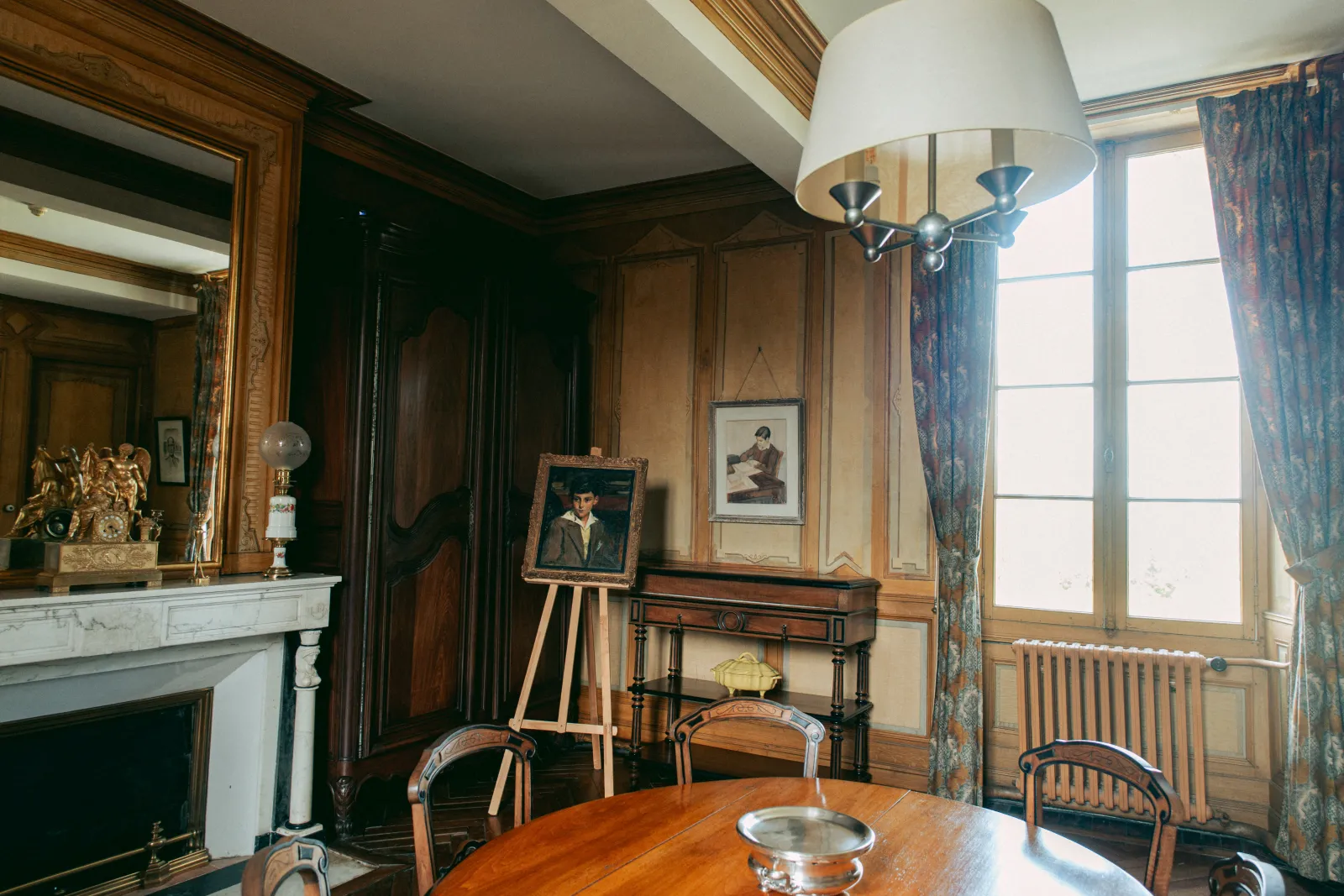 Elegant interior of the Domaine de Malagar in Bordeaux featuring a dining room with round wooden table, antique chairs, white marble fireplace, framed portraits, white chandelier, wood paneling on walls, and large windows with blue curtains.