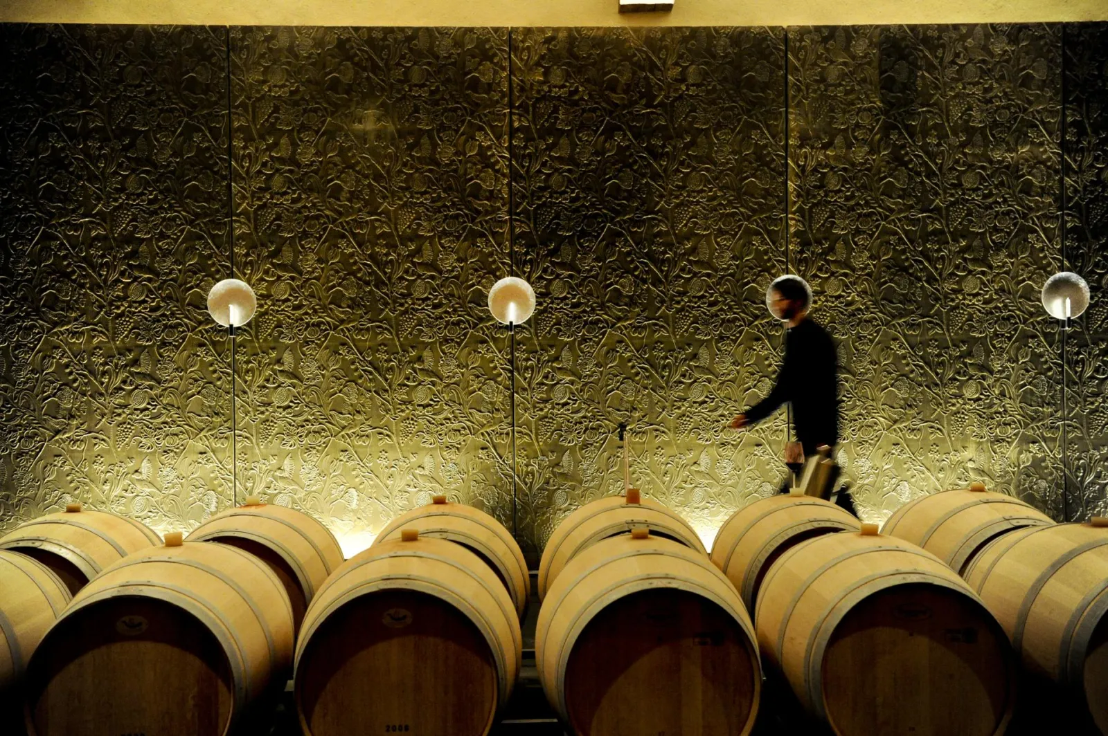 A man in a black suit walks past rows of oak barrels in a Bordeaux château cellar, with an ornate gilded patterned wall and ceiling lights in the background.