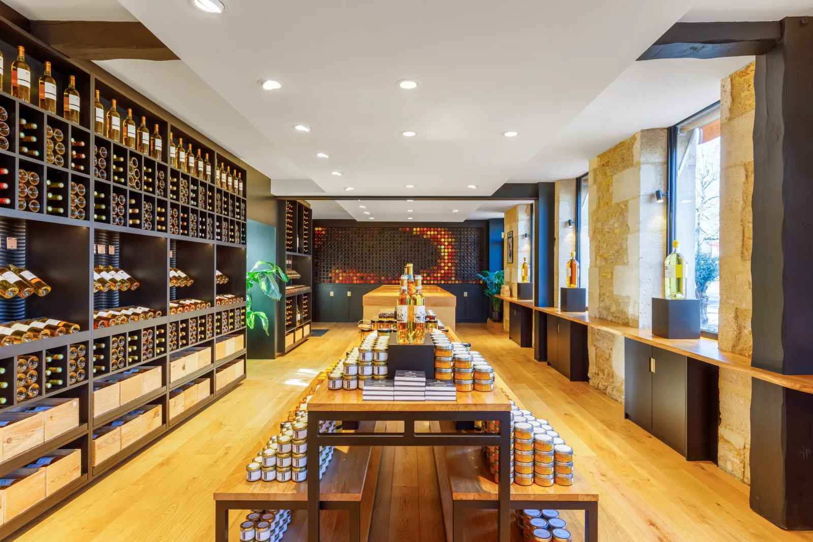 Interior of the Maison du Sauternes in Bordeaux: elongated tasting room with wine bottle shelves, display tables featuring jars, light wooden flooring, modern lighting, and illuminated central showcase.
