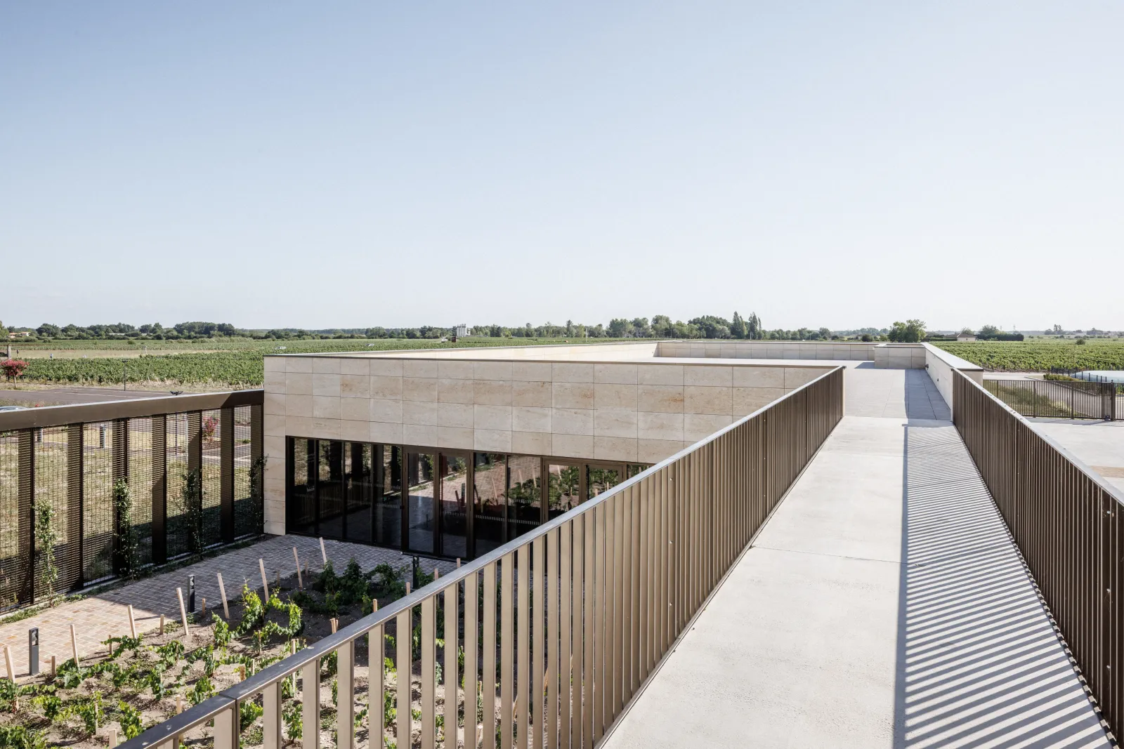 Aerial view of a modern concrete building with flat roof surrounded by vineyards, accessed by a metal walkway with railings. Château Larose-Trintaudon in Bordeaux.