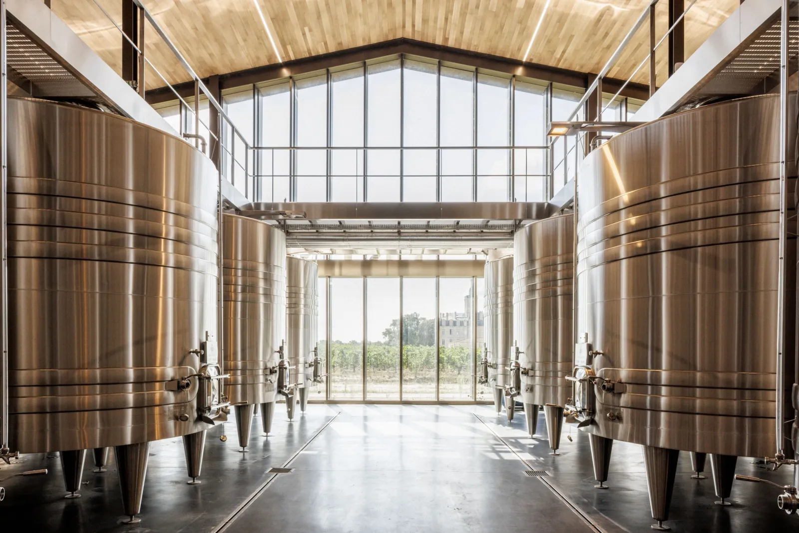 Modern winery interior featuring stainless steel fermentation tanks lined on both sides, large windows overlooking Bordeaux vineyards, contemporary architecture with vaulted ceiling and natural lighting.