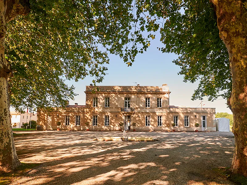 Château Haut Breton Larigaudière in Bordeaux, a 19th-century ochre stone mansion framed by ancient trees, featuring a symmetrical facade and paved courtyard.