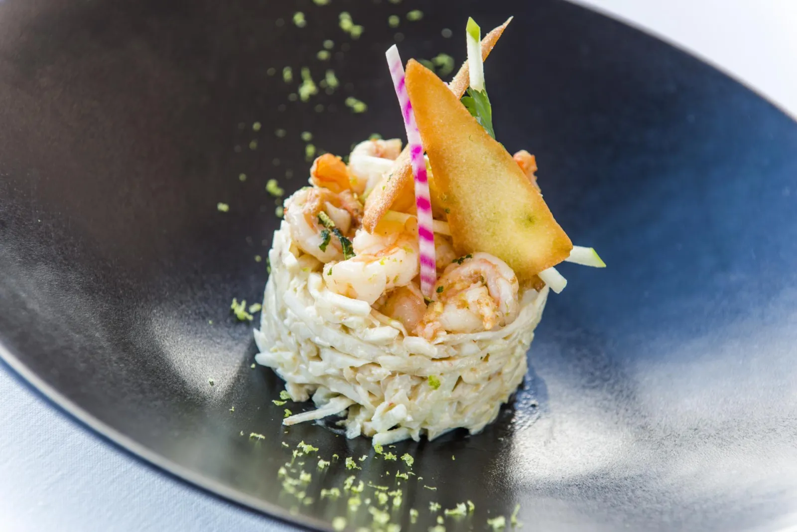 Gourmet dish served on a black plate: nest of thin pasta topped with shrimp, pan-seared pear and colorful garnish, green powder dusting