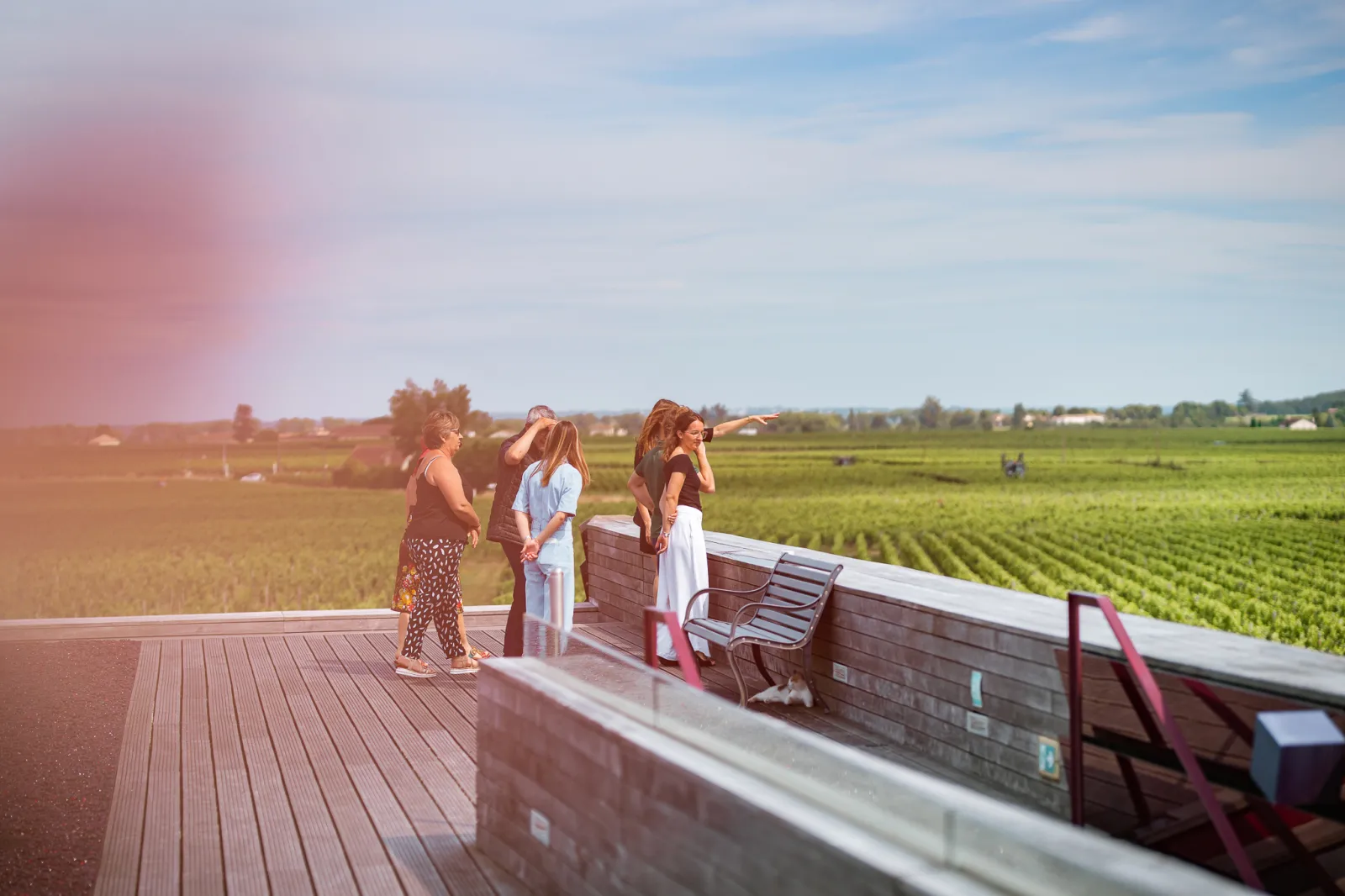 Three women standing on a wooden terrace overlooking the Bordeaux vineyards at Château La Dominique, during a sunny late afternoon visit.