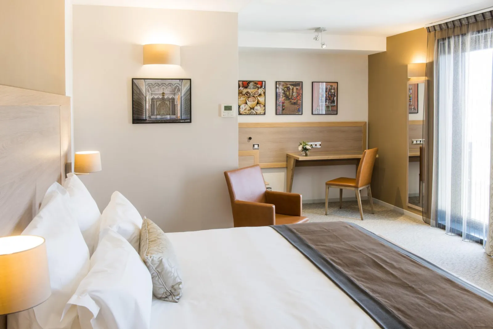 Modern hotel room at Vatel Hotel in Bordeaux featuring a king-size bed, light wood desk, brown armchairs, gold lighting fixtures and contemporary wall art
