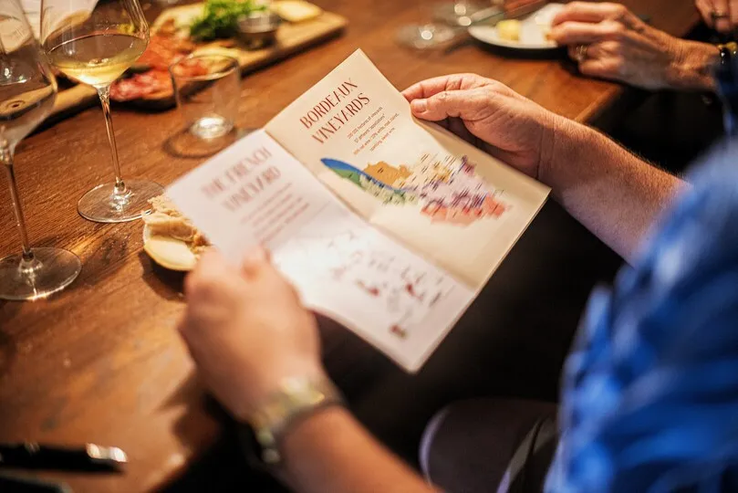 A person holds a Bordeaux Vineyards brochure during a wine tasting, with white wine glasses and plates of cheese and charcuterie on the wooden table