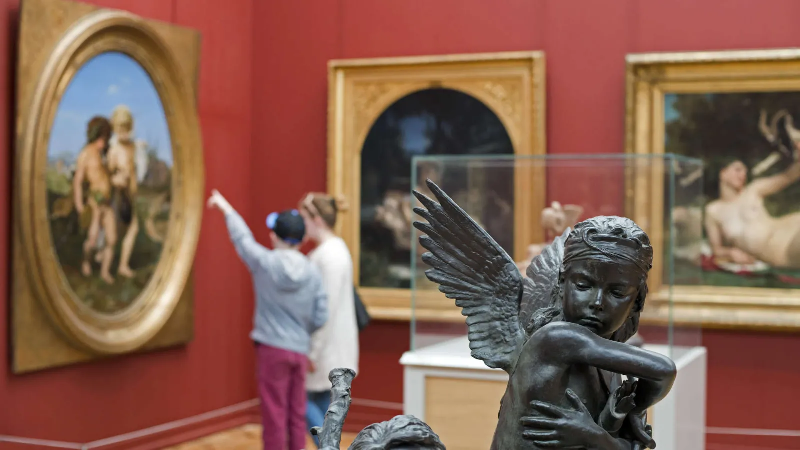 Interior of Bordeaux Museum of Fine Arts with red walls, gilded paintings and sculptures. A visitor photographs an artwork while a bronze angel statue is visible in the foreground.