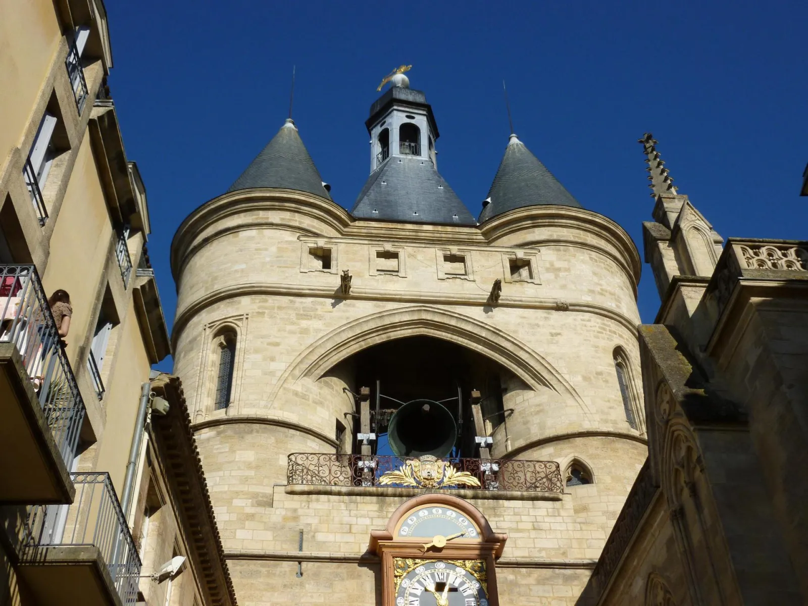 The Grosse Cloche of Bordeaux, a medieval bell tower featuring two slate-roofed turrets, a gilded clock face, and a visible bell, surrounded by historic buildings against a clear blue sky.
