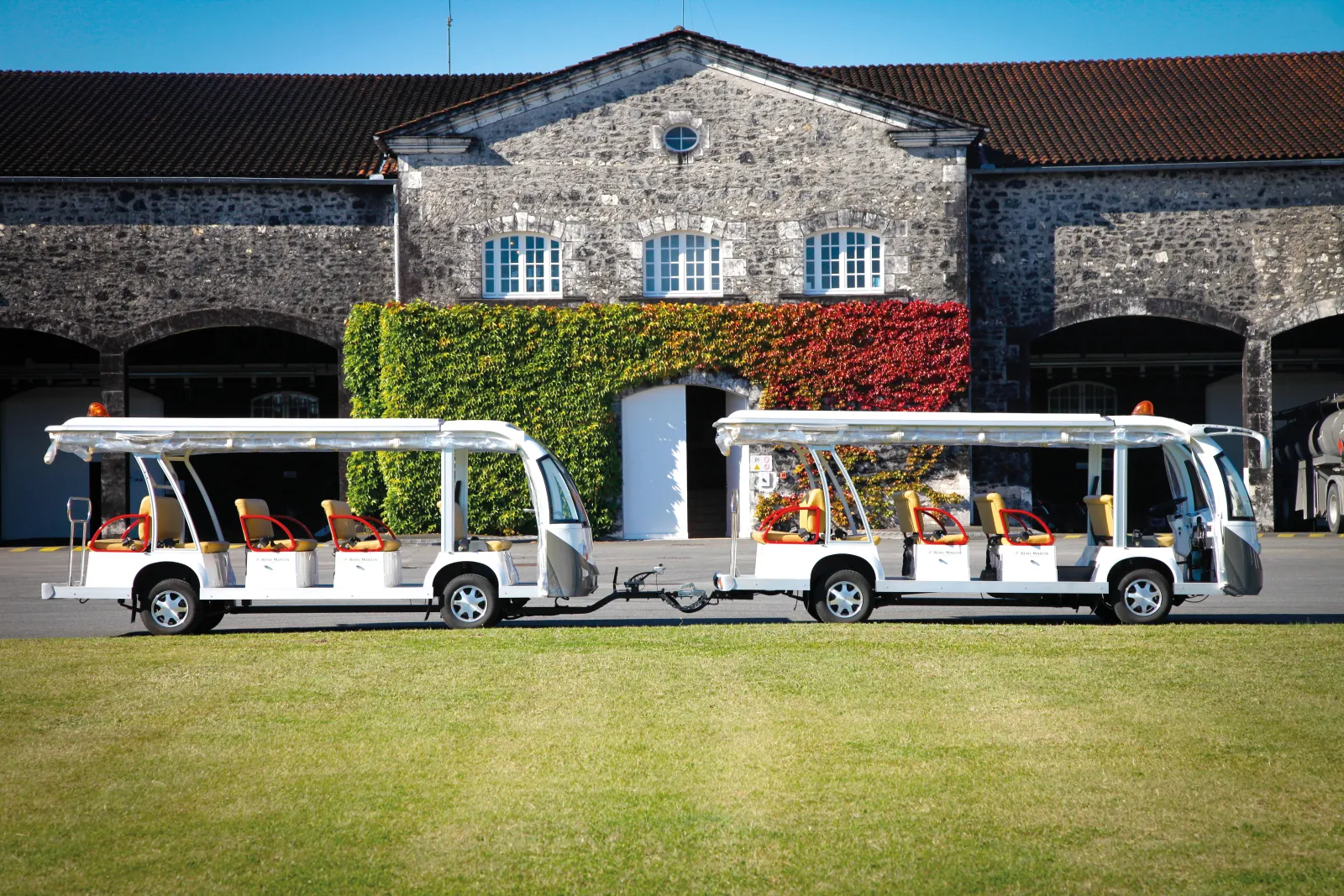 White two-car tourist train parked in front of Rémy Martin house in Bordeaux, featuring stone facades and decorative ivy in the background.