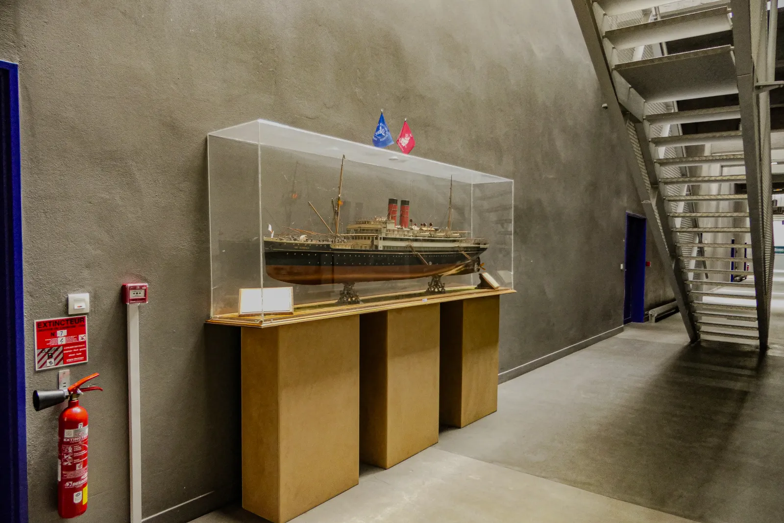 Scale model of an early 20th-century transatlantic steamship displayed in a glass case at the Bordeaux Maritime History Museum, with flags on the base and safety equipment nearby.