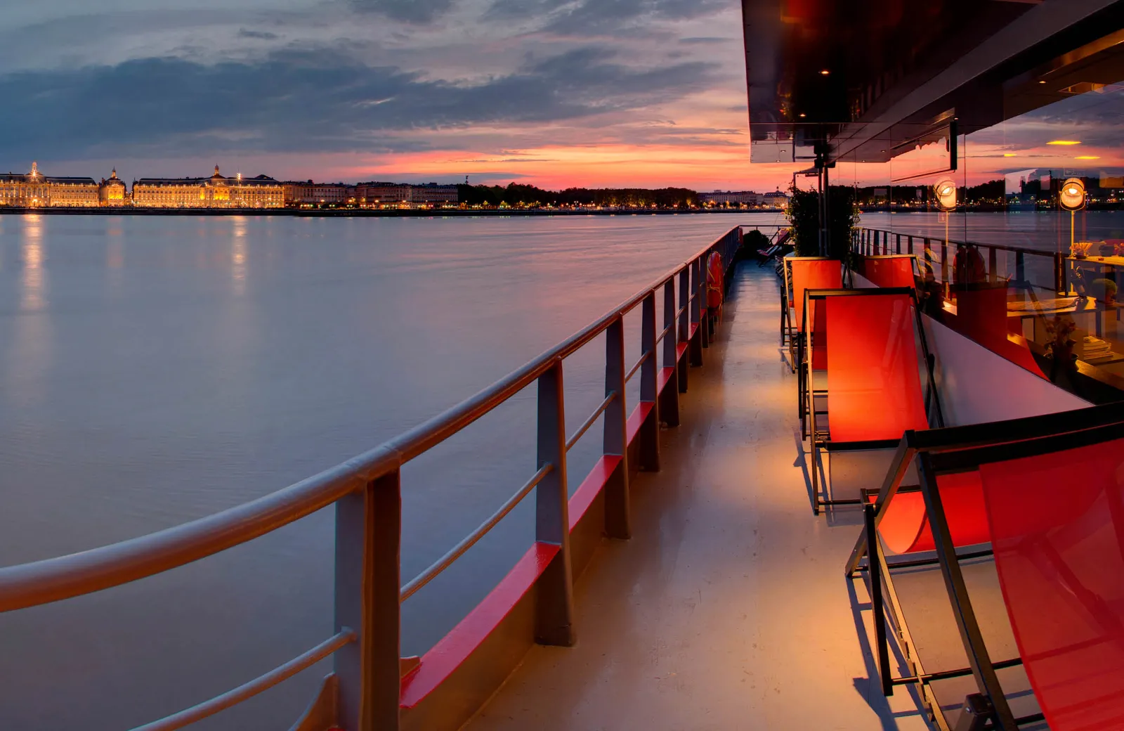 View of the Garonne river from a boat deck at sunset in Bordeaux, with red deck chairs and the illuminated city buildings reflected in the water