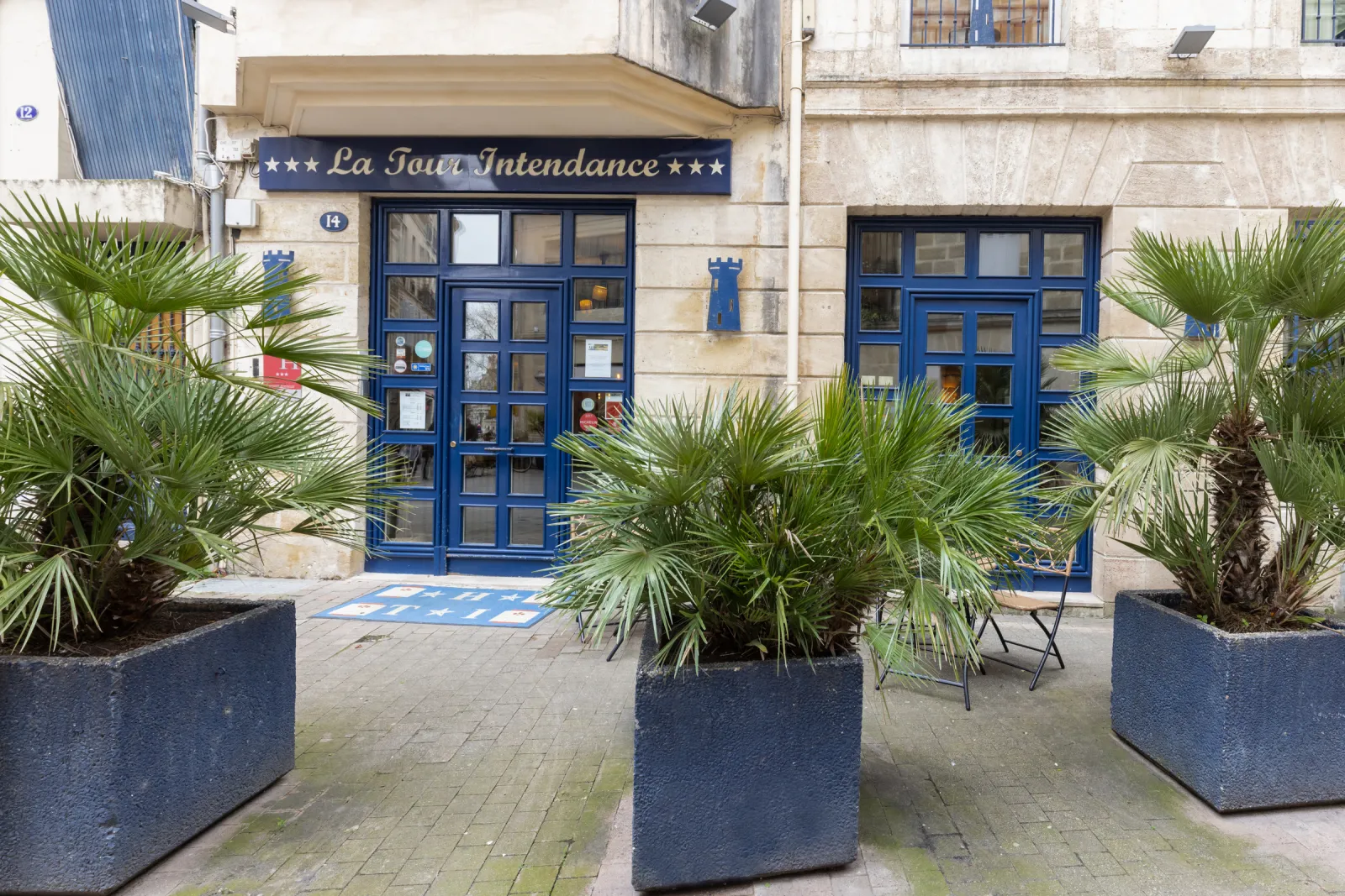 Facade of the Hotel de la Tour Intendance in Bordeaux featuring blue doors, navy signage and potted plants on the paved courtyard