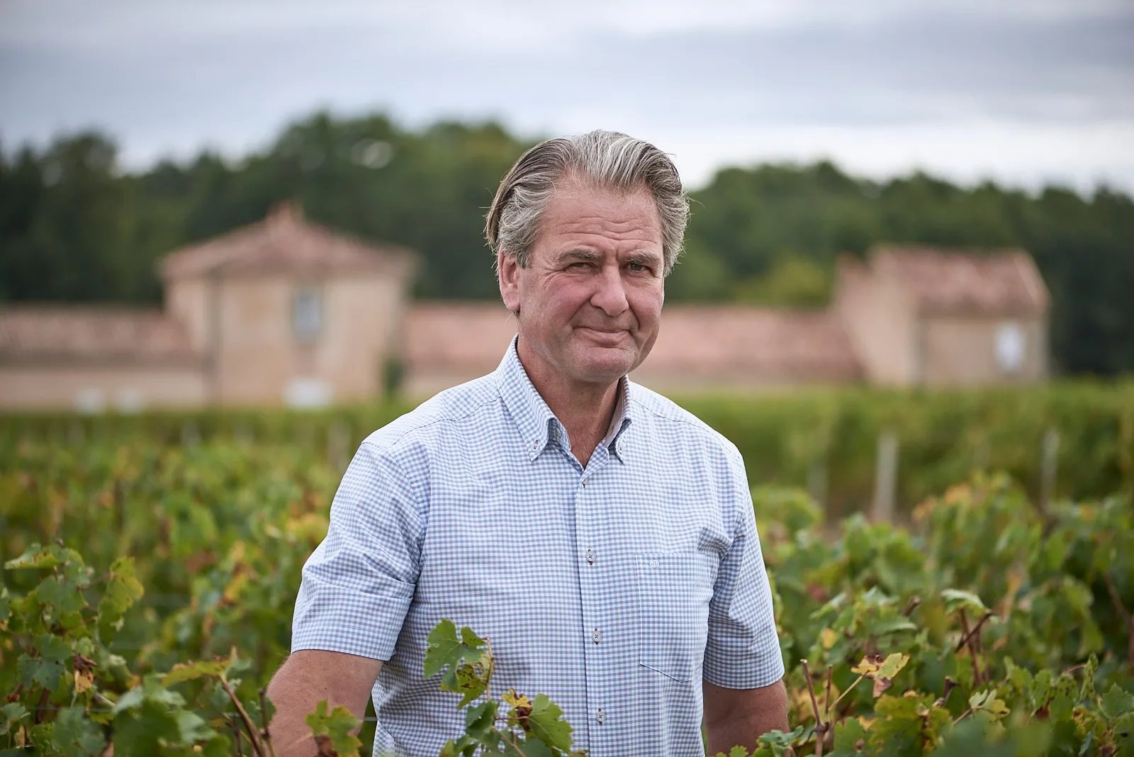 Middle-aged man in light blue checkered shirt standing in a Bordeaux vineyard with Château Desmirail in the background