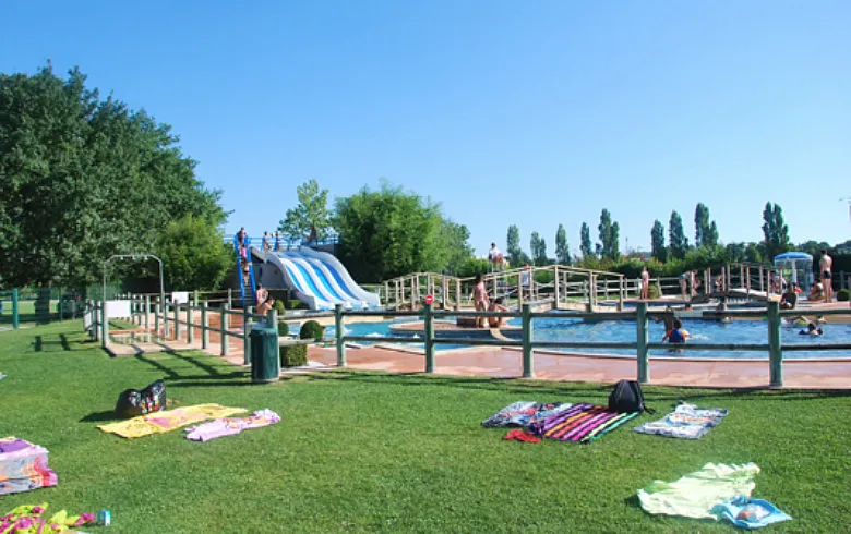 Pinsan swimming pool in Eysines featuring blue water slides, wooden bridges, swimming basins, green lawns, and visitors relaxing on towels.