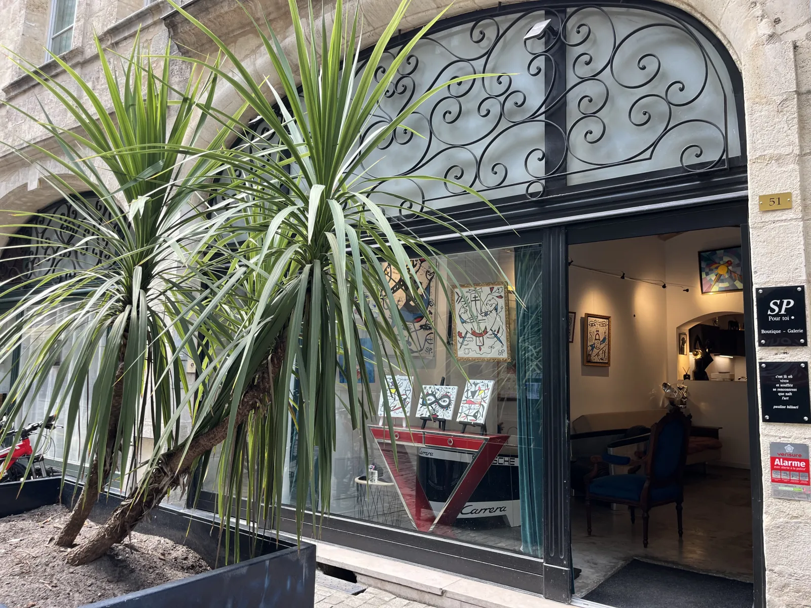 Storefront of SP Pour toi gallery in Bordeaux featuring black glass window, arched door with ornate ironwork, and potted green plant at the entrance