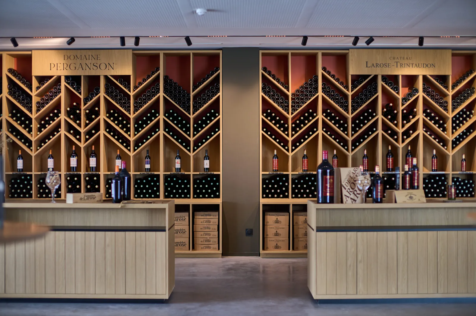 Modern wine tasting cellar displaying bottles from Domaine Perganson and Château Larose-Trintaudon in Bordeaux, stored in illuminated chevron-patterned wooden shelving.
