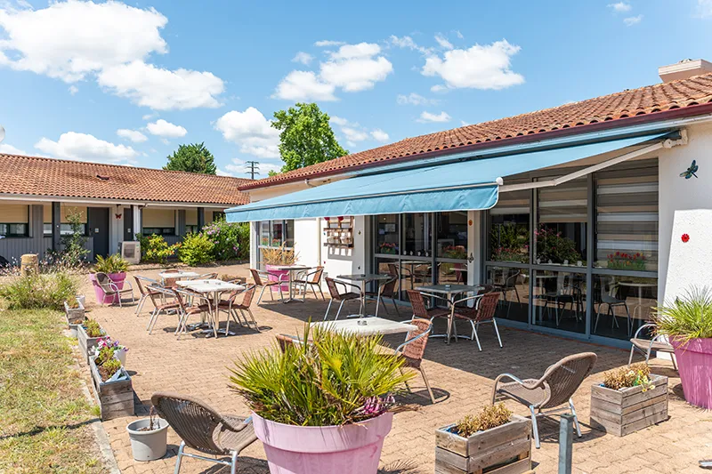 Outdoor patio of Kyriad Direct Bordeaux Sud Cestas hotel featuring tables, pink chairs, flowering planters, and blue awning on a sunny day