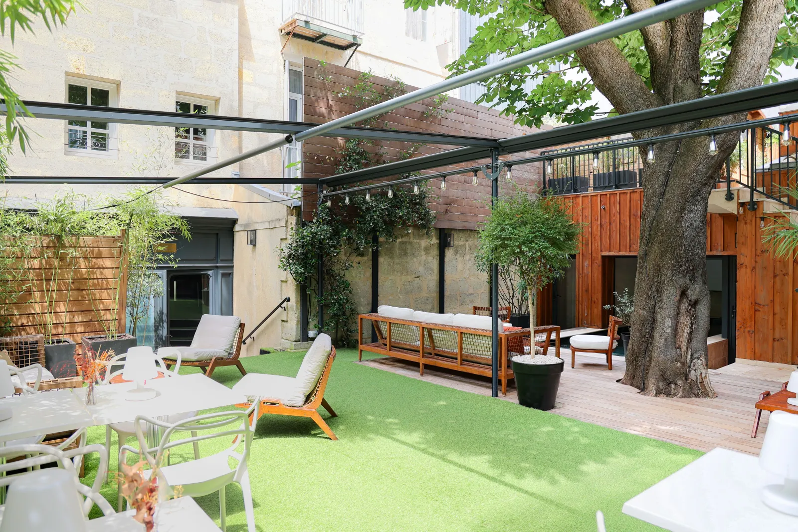 Interior courtyard of a Bordeaux boutique hotel featuring green lawn, wooden lounge furniture, landscape design, modern pergola, and historic buildings