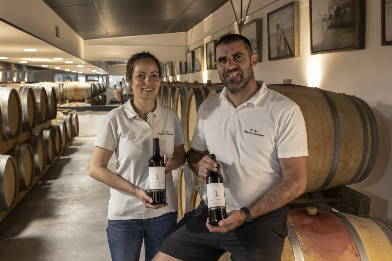 Two winemakers from Château Pierre de Montignac in Bordeaux pose with wine bottles in the cellar surrounded by oak barrels.