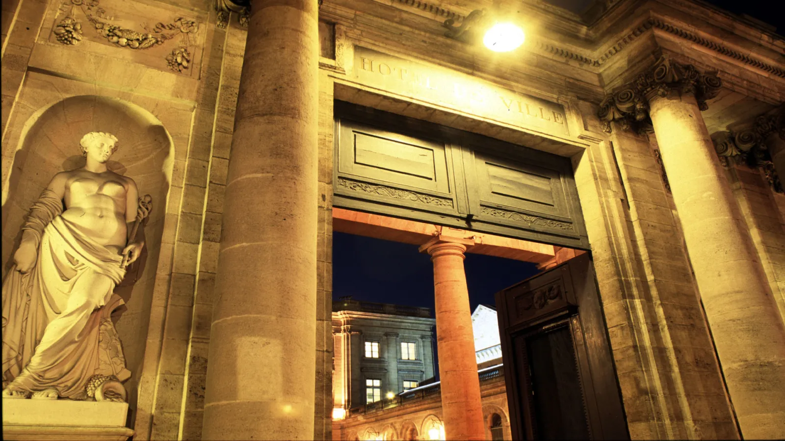 Night view of the Palais Rohan in Bordeaux featuring classical stone sculpture in an arched niche, ornate columns, and illuminated entrance door
