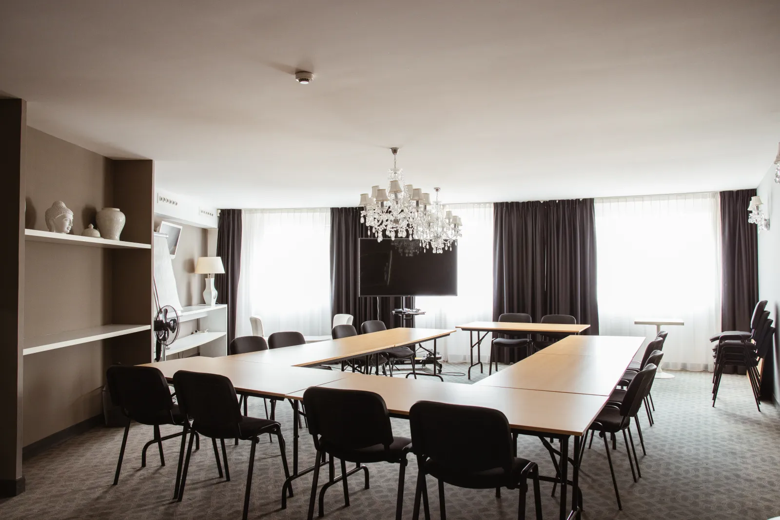 Modern meeting room at Ténéo Apparthôtel Talence Arthena in Bordeaux, featuring white tables, black chairs, crystal chandelier, gray curtains, and large windows with outdoor views.