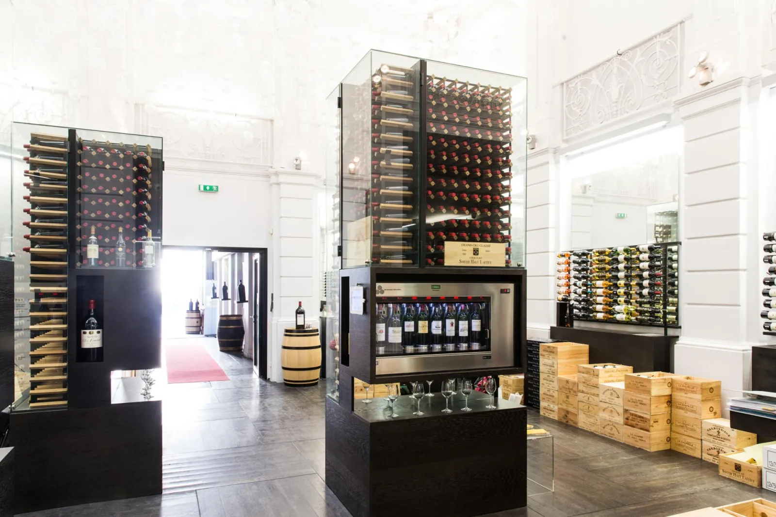 Contemporary interior of Max Bordeaux Wine Gallery & Cellar featuring illuminated wine displays with bottles in glass cases, stacked wooden wine crates, and tasting equipment in a bright, minimalist space.