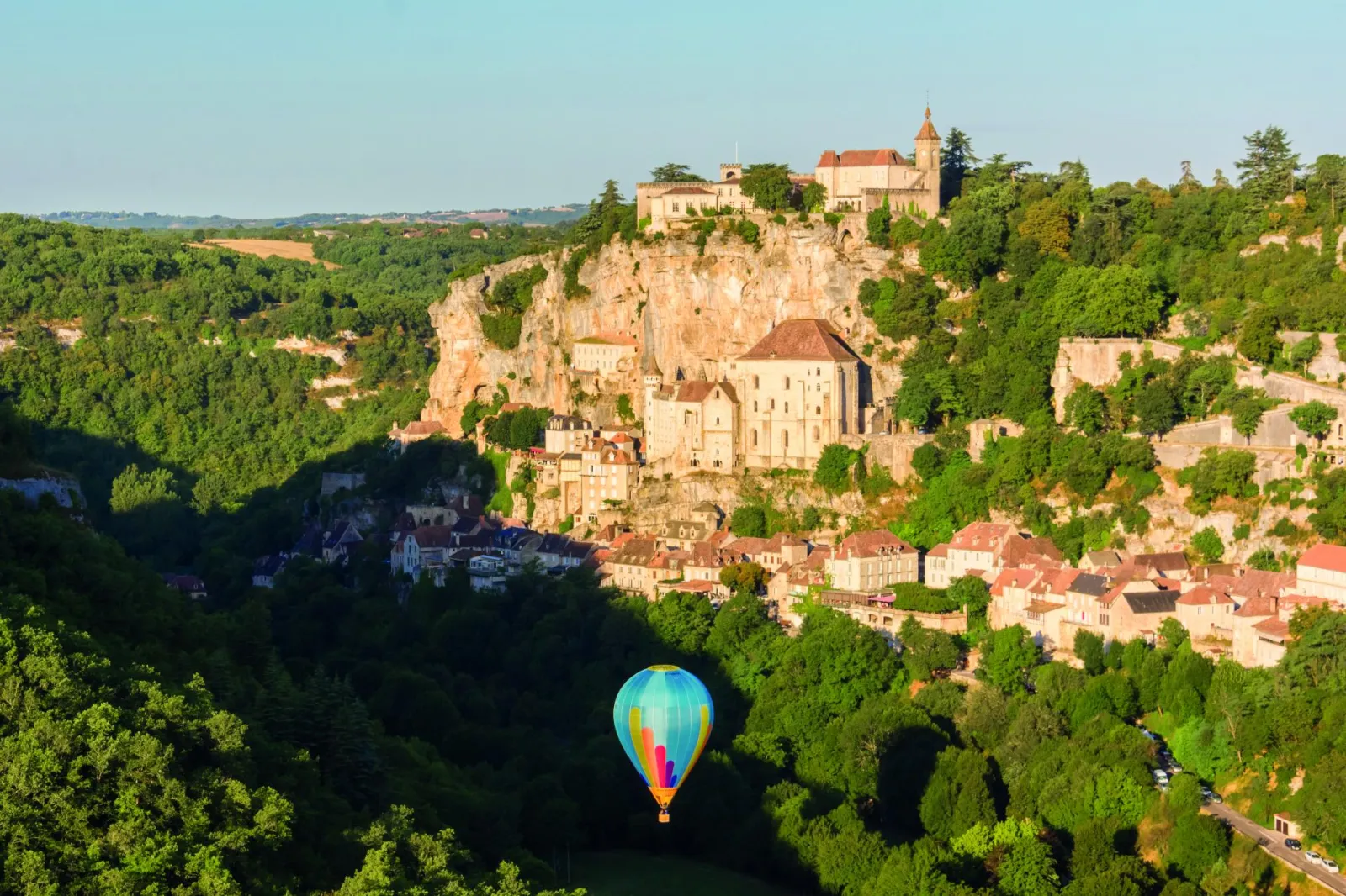 A colorful hot air balloon flying over a forested valley with the medieval village of Rocamadour perched on a limestone cliff, showcasing its castle and stone churches.