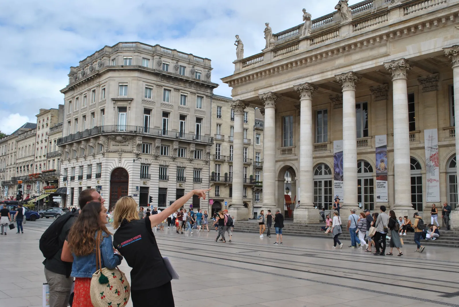 Two tourists admiring the Place de la Comédie in Bordeaux, with the majestic Grand Theatre and its impressive columns in the background, surrounded by visitors and Haussmann-style buildings.