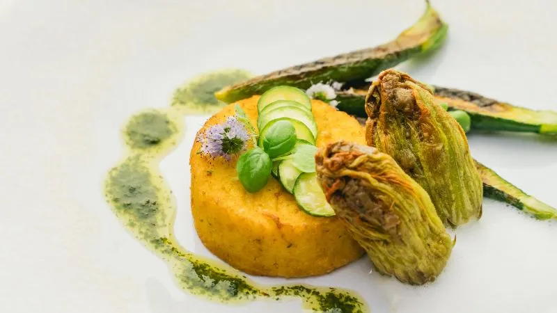Gourmet plated dish on white plate: yellow-orange stuffed tomato topped with thin cucumber slices and green sprouts, accompanied by stuffed zucchini blossoms and drizzled green sauce.