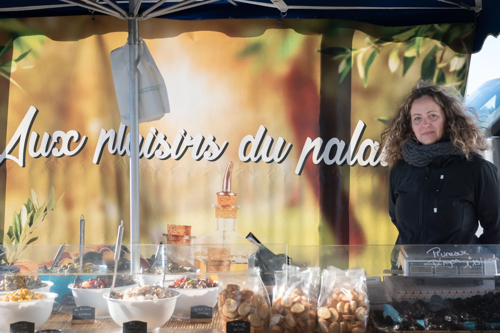 Woman at a market stall in Bouliac, Bordeaux, displaying food products under a blue parasol with 'Halle Palissy du Palais' signage, surrounded by bowls and jars of culinary specialties.