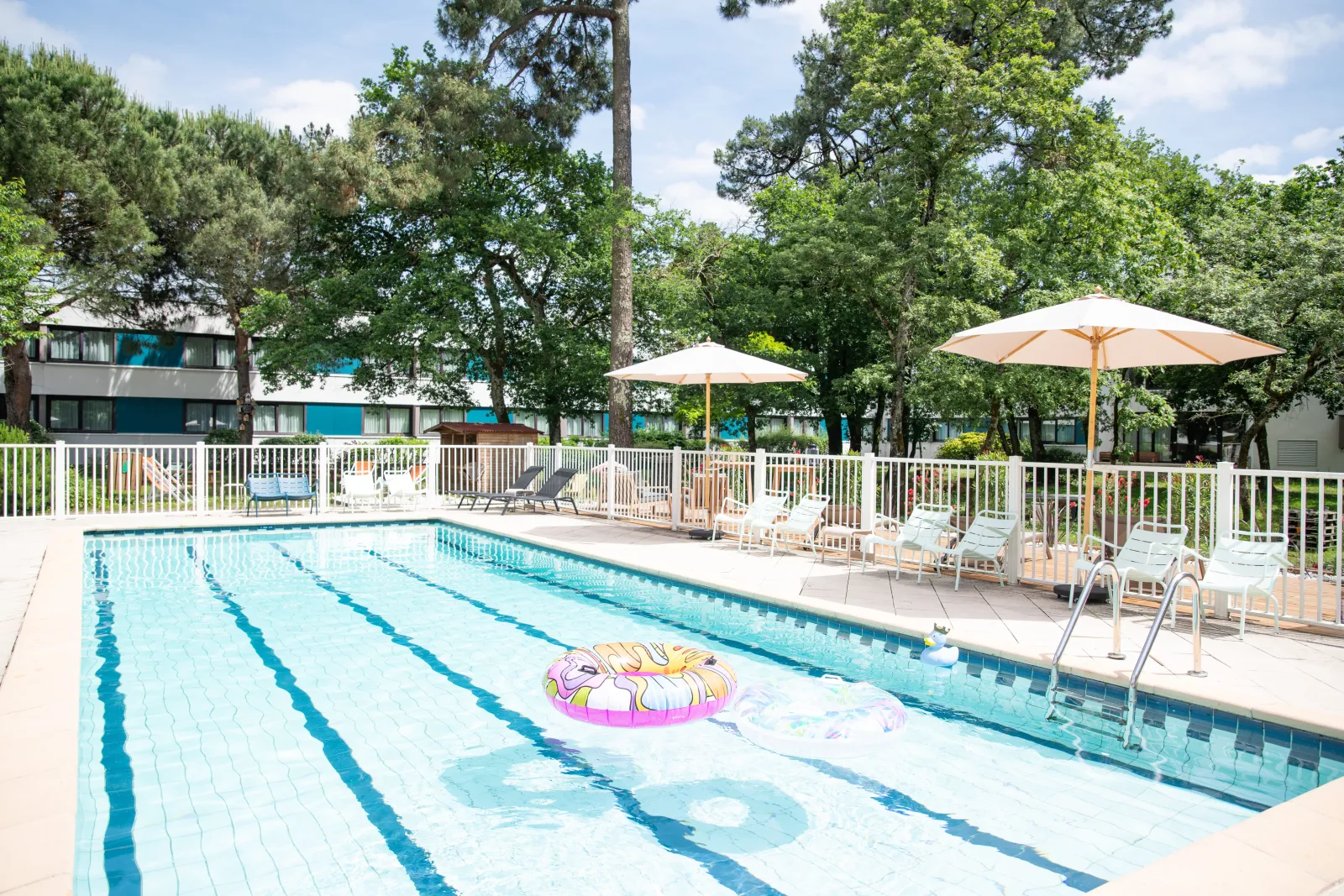 Olympic swimming pool with lap lanes at Novotel Bordeaux Mérignac hotel, surrounded by loungers, white and beige umbrellas, white fence, hotel building and trees in the background